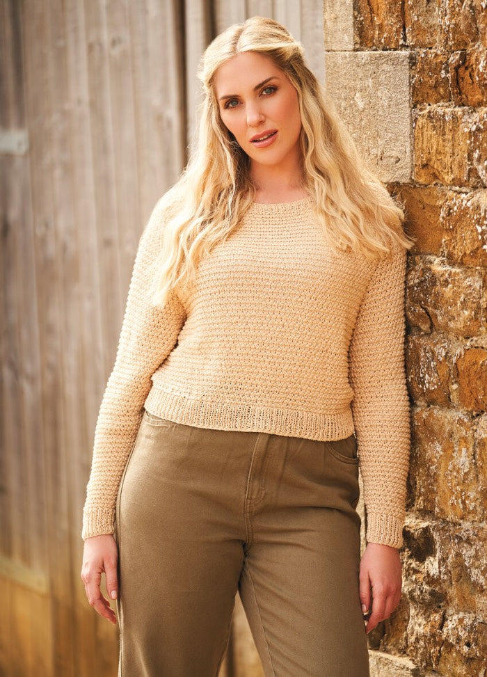 A woman in a beige sweater and brown pants leans against a rustic stone wall, exuding a relaxed and confident demeanor. Wooden siding adds a cozy, natural backdrop.