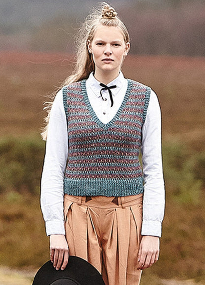 Person wearing the Fell Vest, a striped knit in blue and brown with V-neck, styled over a white shirt and pleated pants. Photographed in a natural landscape.