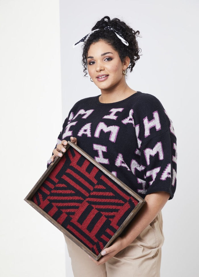 A woman stands holding a wooden tray with a red and black geometric pattern. She wears a black sweater with "I AM" in bold light-colored text, exuding confidence.