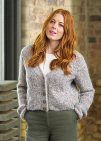 A woman with long red hair stands confidently, wearing a gray knit cardigan and olive pants, set against a rustic brick wall backdrop.