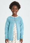 A smiling young girl in a light blue knitted cardigan stands against a plain background. She wears a cream dress with small rainbow patterns, exuding a joyful and cheerful tone.