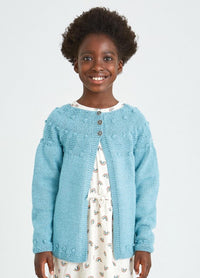 A smiling young girl in a light blue knitted cardigan stands against a plain background. She wears a cream dress with small rainbow patterns, exuding a joyful and cheerful tone.