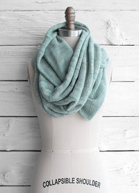 Fino Circle Scarf displayed on a collapsible shoulder dress form, featuring a light blue knitted design elegantly draped around the neck, set against a white wooden background.