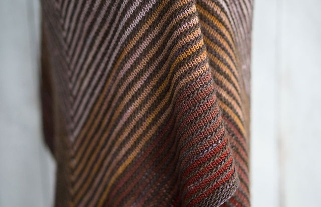 Fino Shadow Shawl on a mannequin, with gradient stripes in brown, orange, and red forming a V-shape, shown against a wooden background.
