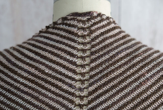 Fino Shadow Shawl on a mannequin, with gradient V-shaped stripes in brown, orange, and red, set against a wooden background.