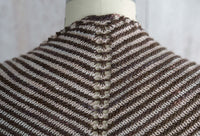 Fino Shadow Shawl on a mannequin, with gradient V-shaped stripes in brown, orange, and red, set against a wooden background.
