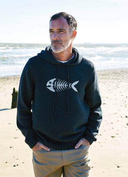 A man in a dark sweater with a fish skeleton design stands on a sandy beach, hands in pockets. The ocean waves calmly in the background under a clear sky.
