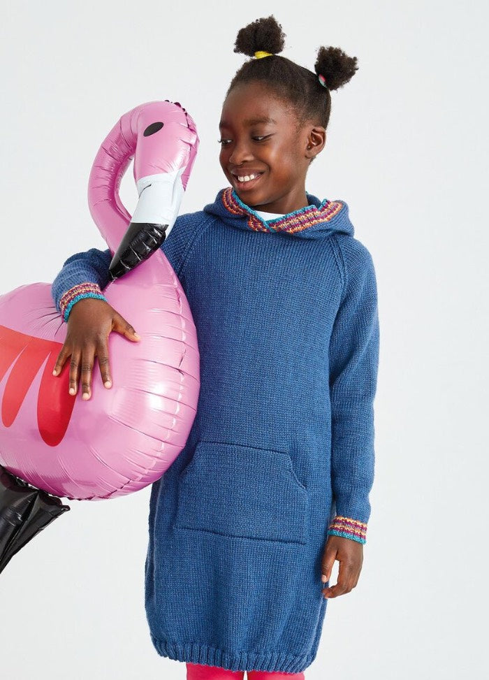 A young girl smiles while holding a pink flamingo balloon. She wears a blue sweater dress with a front pocket. The mood is joyful and playful.