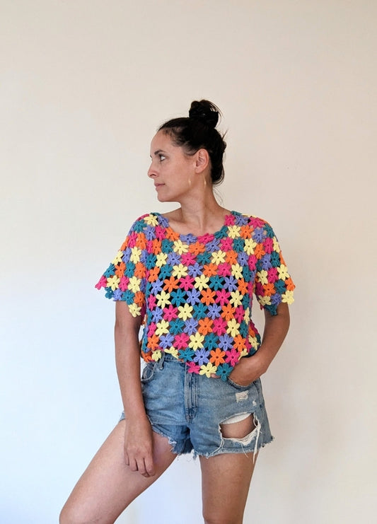 Woman in colourful crochet top and ripped denim shorts looks to the side against a plain background. The mood is casual and vibrant.