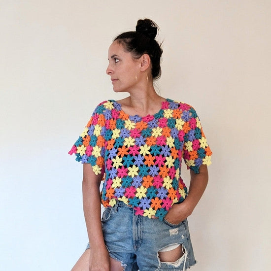 Woman in colourful crochet top and ripped denim shorts looks to the side against a plain background. The mood is casual and vibrant.