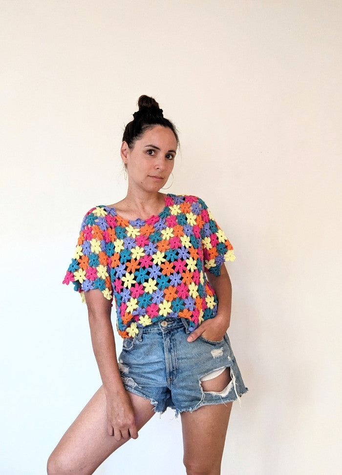 A person poses confidently against a plain light background, wearing a colourful crochet top and distressed denim shorts. The mood is casual and vibrant.
