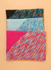 Stacked Florence Blankets in black, light blue, and bright pink, each with distinct multicolored patterns and trims.