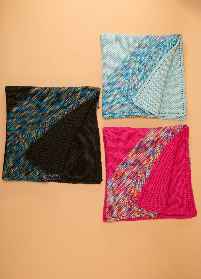 Folded Florence Blankets in light blue, black, and bright pink, each showing a patterned side, arranged on a light orange surface.