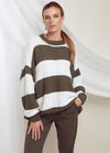 A woman stands confidently wearing a loose, striped sweater in olive green and white. She pairs it with olive trousers, exuding a casual, relaxed vibe.
