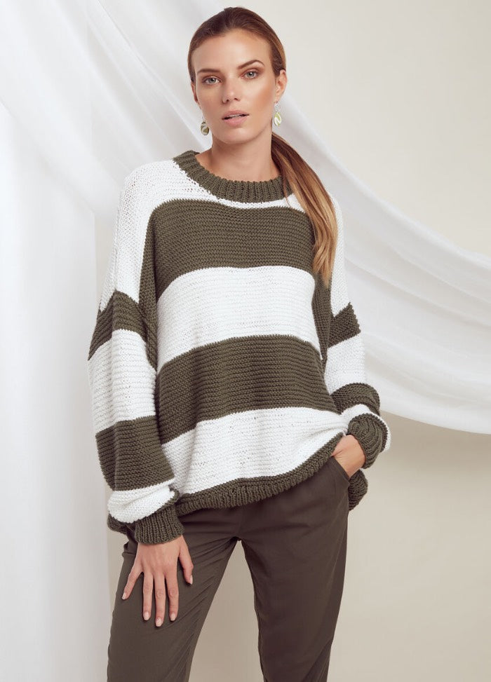 A woman stands confidently wearing a loose, striped sweater in olive green and white. She pairs it with olive trousers, exuding a casual, relaxed vibe.