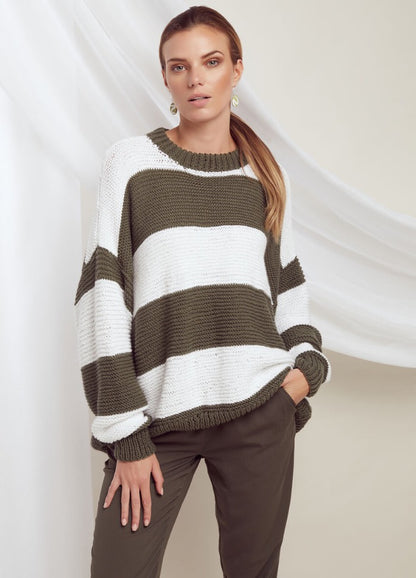 A woman stands confidently wearing a loose, striped sweater in olive green and white. She pairs it with olive trousers, exuding a casual, relaxed vibe.