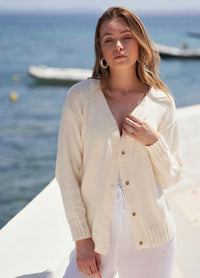 A woman stands by the seaside wearing a light cream cardigan and white pants. The ocean and a boat are visible in the background, creating a serene and relaxed atmosphere.