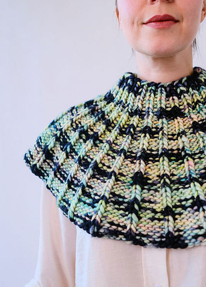 Model wearing the Franca Shoulder Cozy, a ribbed knit in chunky black, green, yellow, and pink yarn, covering shoulders and upper chest.