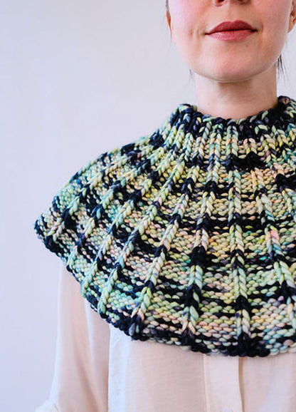 Model wearing the Franca Shoulder Cozy, a ribbed knit in chunky black, green, yellow, and pink yarn, covering shoulders and upper chest.