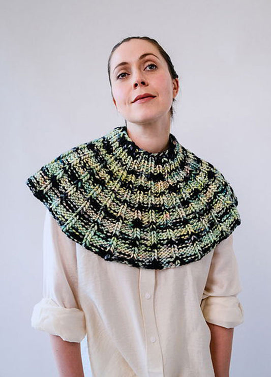Model wearing the Franca Shoulder Cozy, a ribbed knit in chunky green, black, and beige yarn, styled over a light shirt.