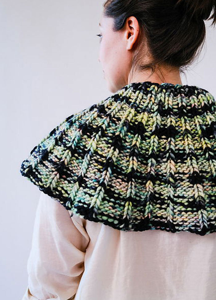 Model wearing the Franca Shoulder Cozy, a chunky ribbed knit in black, green, and beige, styled over a light button-up shirt.