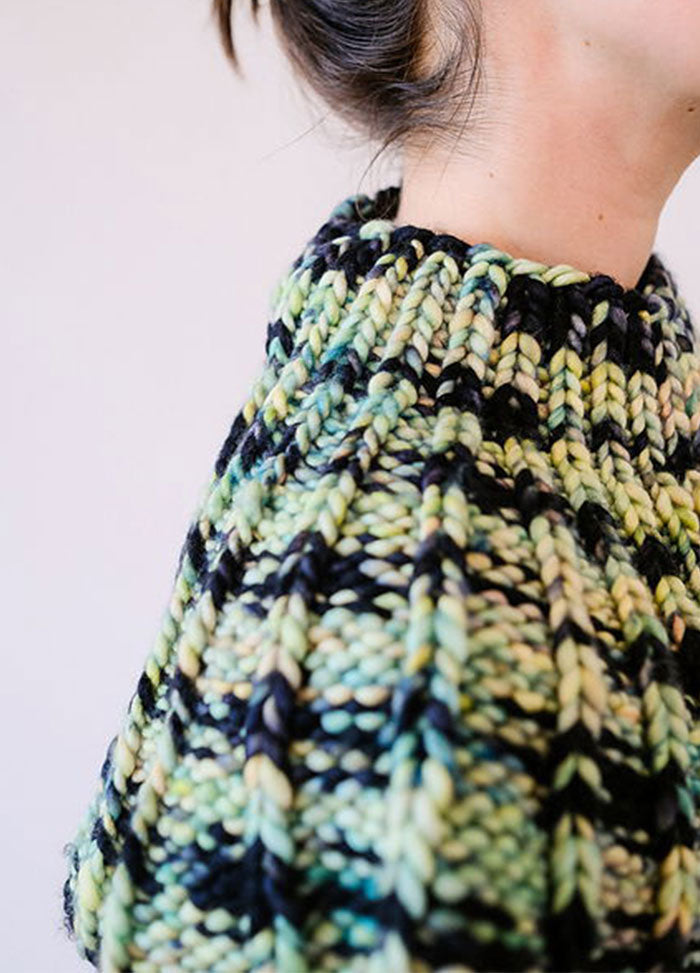Model wearing the Franca Shoulder Cozy, a chunky ribbed knit in black, green, and beige, styled over a light button-up shirt.