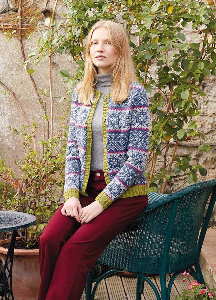 A woman sits on a teal chair in a garden, wearing a colorful knit cardigan, gray top, and red pants, conveying a calm and cozy atmosphere.