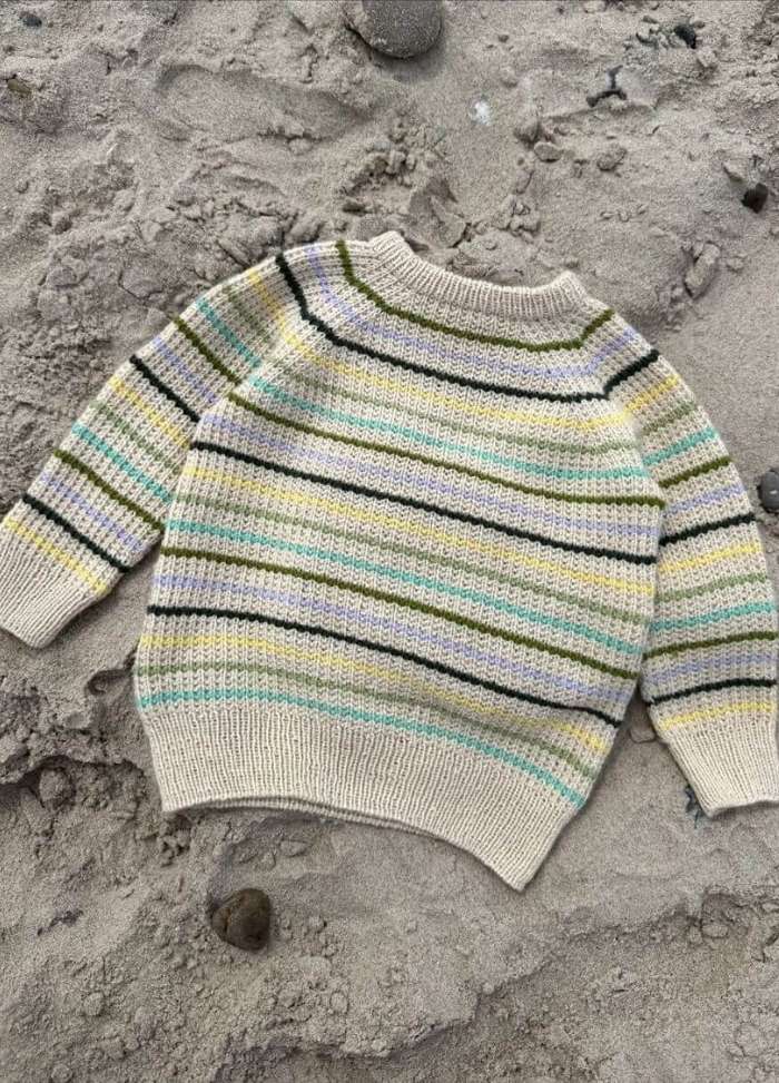 A cozy, multicolored striped sweater with light yellow, green, and black stripes lies on sand. The sweater evokes a casual and warm vibe.