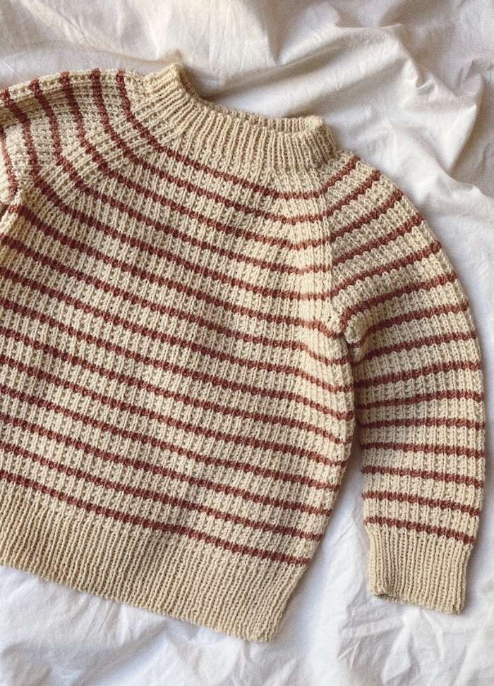 Cozy knitted sweater with beige and brown stripes laid on crumpled white fabric. The garment has a ribbed neck, cuffs, and hem, evoking warmth.