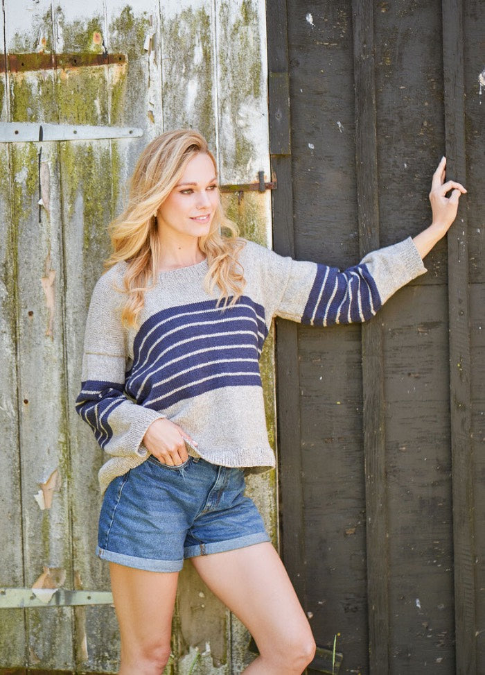 A woman in a striped sweater and denim shorts leans against a rustic wooden wall. She looks to the side, exuding a relaxed, contemplative vibe.