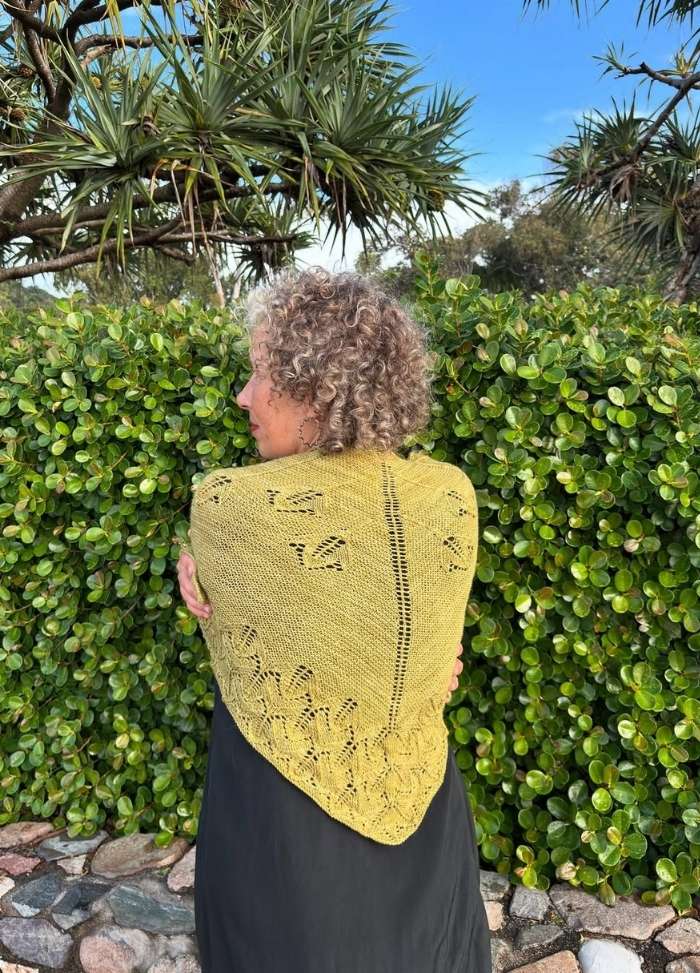 Curly-haired woman wearing a yellow knitted shawl with floral patterns stands against lush green foliage and bright blue sky, evoking tranquility.