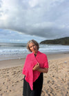 A woman with curly hair wearing a bright pink shawl stands on a sandy beach with waves and a cloudy sky in the background, exuding a serene mood.