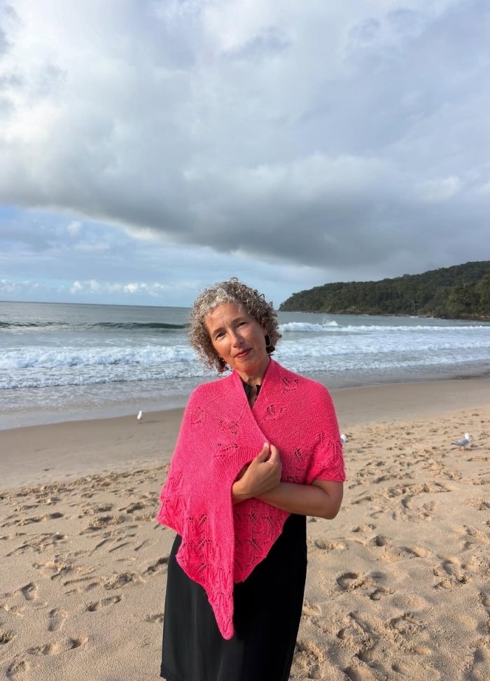 A woman with curly hair wearing a bright pink shawl stands on a sandy beach with waves and a cloudy sky in the background, exuding a serene mood.