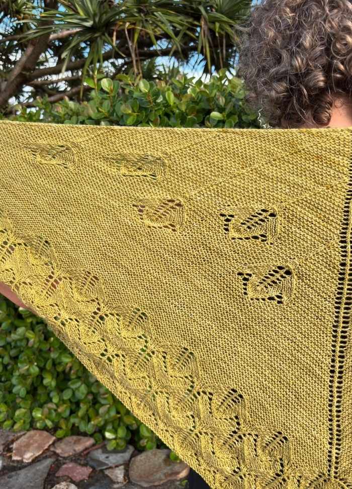 A person holds a yellow knitted shawl with intricate leaf and bird patterns, against a background of lush greenery. The tone is peaceful and natural.