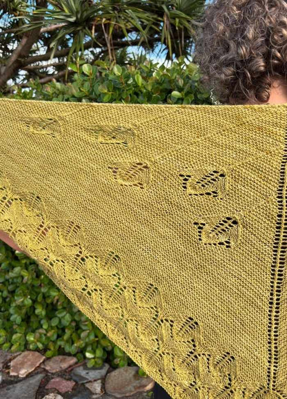 A person holds a yellow knitted shawl with intricate leaf and bird patterns, against a background of lush greenery. The tone is peaceful and natural.