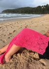 Person relaxing on a sandy beach with waves in the background. A vibrant pink lace cover drapes their legs, under a cloudy sky. Tranquil mood.