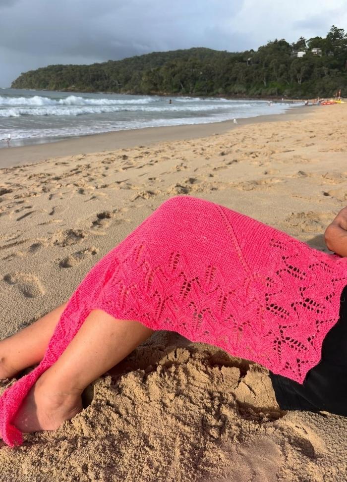 Person relaxing on a sandy beach with waves in the background. A vibrant pink lace cover drapes their legs, under a cloudy sky. Tranquil mood.