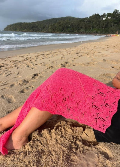 Person relaxing on a sandy beach with waves in the background. A vibrant pink lace cover drapes their legs, under a cloudy sky. Tranquil mood.