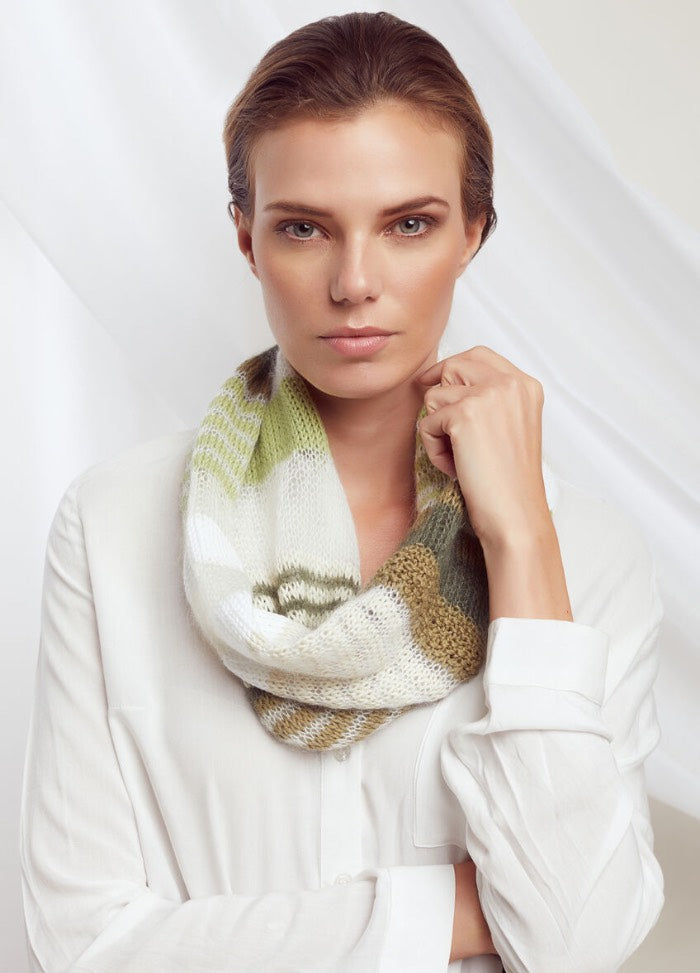 A person with a calm expression wears a white blouse and a multicolored knitted scarf in earthy tones. The background is a soft, light fabric.