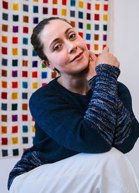 Model in a navy hand-knit Galaxy Sweater with white and blue speckles, starry night effect, against a colorful tiled background.