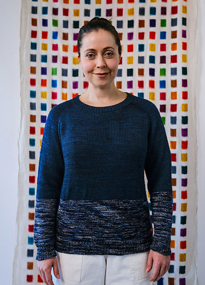 Hand-knit Galaxy Sweater in deep blue with light speckles on lower body and sleeves, shown on a model before a vibrant grid backdrop.