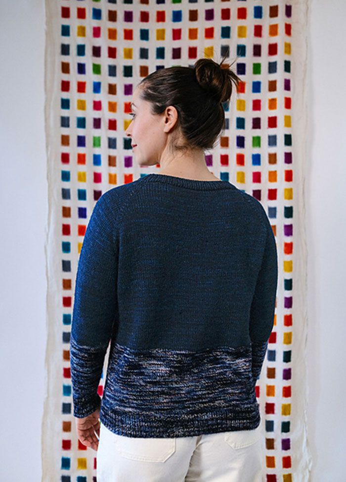 Navy Galaxy Sweater with gradient white and blue speckles on the lower half, modeled in front of a multicolored grid-patterned textile.