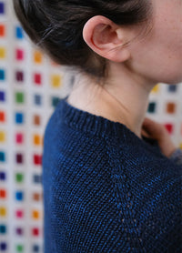 Model wearing a dark blue Galaxy Sweater with galaxy-inspired speckles on the lower body and sleeves, set against a colorful grid background.