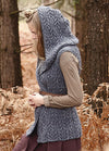 Person wearing the Galloway Vest, a gray hooded cable-knit design over a long-sleeved shirt, photographed outdoors among trees and dried foliage.