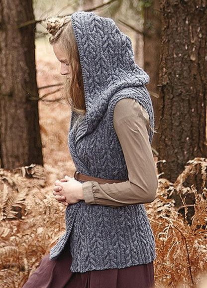 Person wearing the Galloway Vest, a gray hooded cable-knit design over a long-sleeved shirt, photographed outdoors among trees and dried foliage.