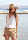 Young woman wearing straw hat, white sleeveless top, and tan shorts gazes thoughtfully by the seaside. Bright, relaxed summer day with calm waters.