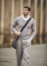 Man walking in an urban setting, wearing a light gray sweater over a white shirt, beige pants, and carrying a shoulder bag. He looks pensive and stylish.