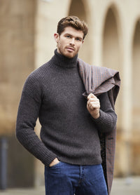 A man stands outside, wearing a gray turtleneck sweater and jeans, with a plaid jacket draped over his shoulder. The scene is relaxed and stylish.