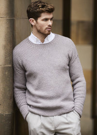 A man with short brown hair stands against a beige wall, wearing a light gray sweater over a white shirt. He looks to the side thoughtfully.