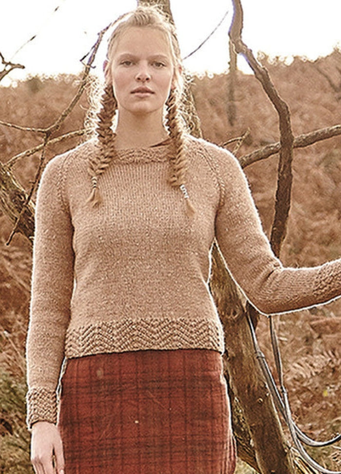 Person wearing the Gotland Sweater, a beige knit with textured cuffs and hem, paired with a red plaid skirt in an autumnal outdoor setting.